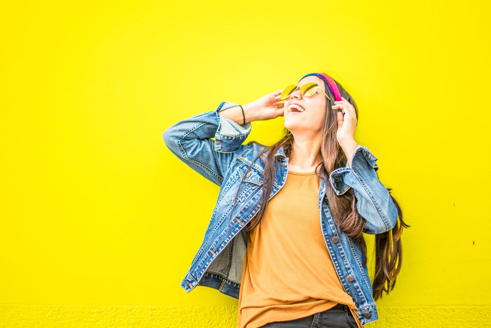 Mulher rindo encostada numa parede amarela chapada, de jaqueta jeans, óculos escuros e tiara colorida