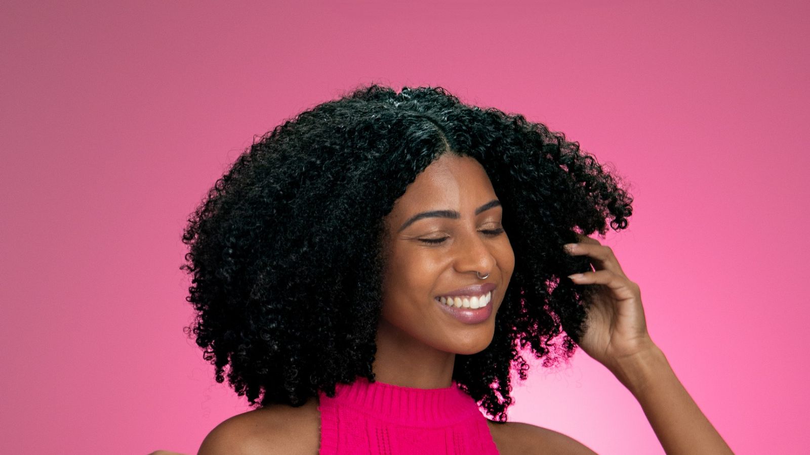 Mulher de cabelo cacheado rindo de blusa pink em frente a uma parede rosa chapada, retrato de estúdio